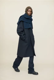 Model wearing dark coat, blue scarf, and brown hood standing against a seamless neutral background