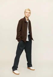 Model with short blonde hair wearing a brown collared jacket, dark relaxed trousers, and white sneakers against a pale backdrop.