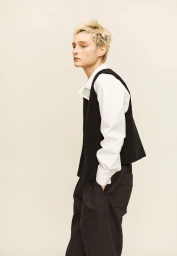 Model with short blonde hair standing in profile wearing a black vest over a white collared shirt and black trousers.