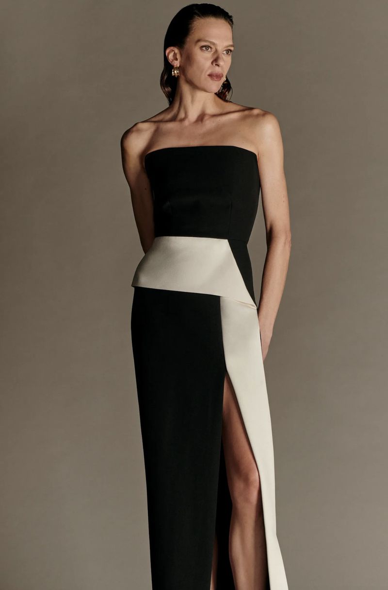 Elegant adult woman in strapless black and white gown with thigh-high slit posing in a studio setting against a grey backdrop.