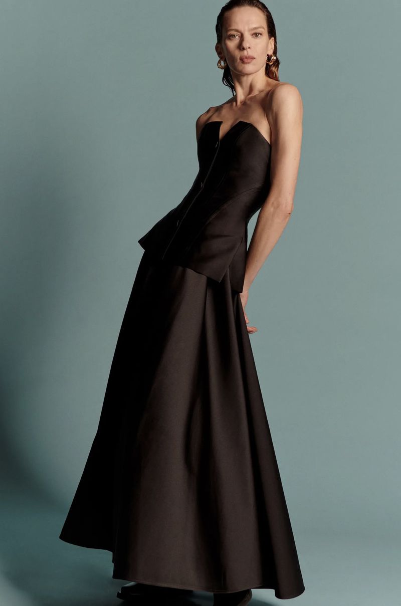 Adult woman poses in black strapless top and skirt against a blue background in a fashion editorial style photograph. 