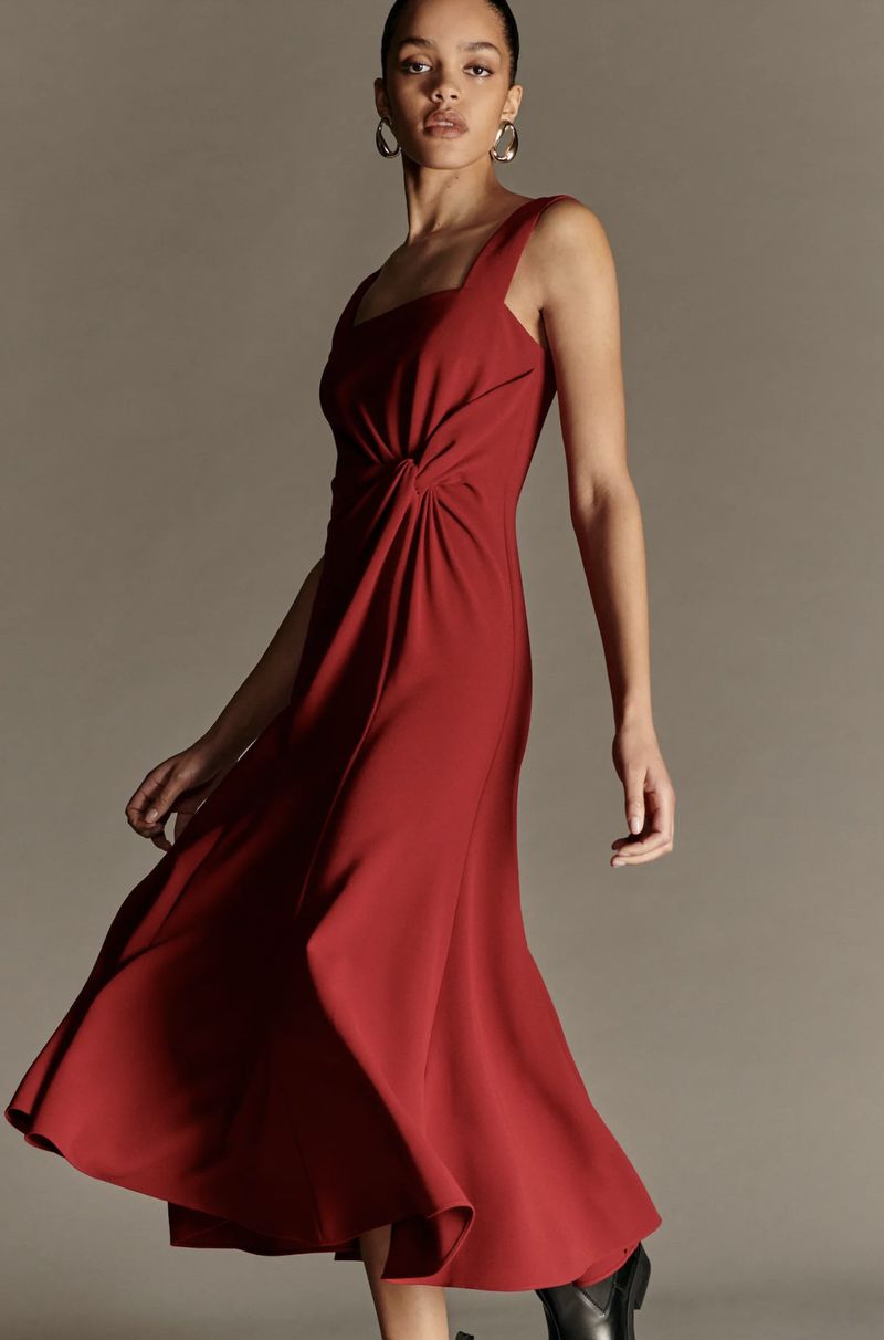 Elegant woman in a red midi dress with a fitted bodice, posing in a studio with a neutral background. She wears hoop earrings.