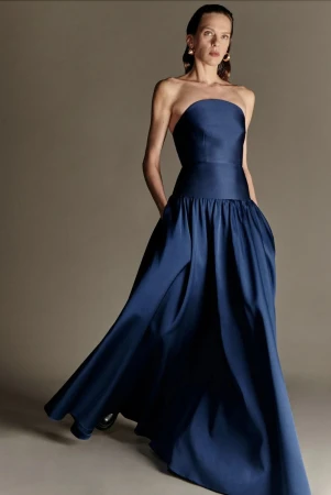 Model wearing a strapless navy blue evening gown with a drop waist and full pleated skirt against a studio backdrop.