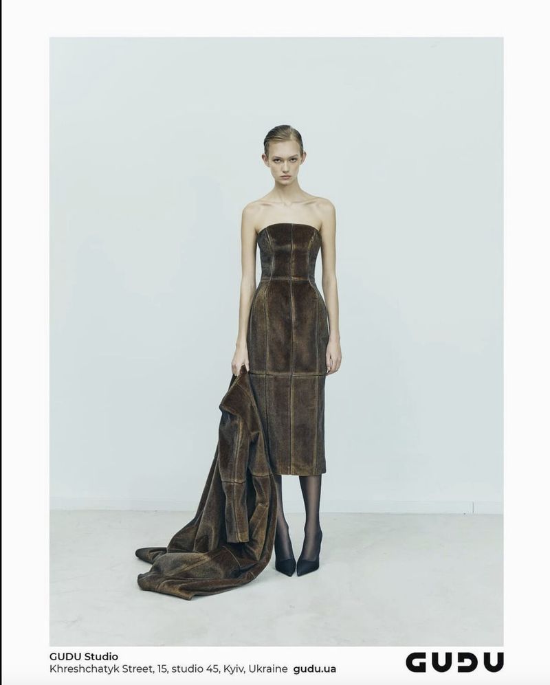 A woman posing in a brown strapless dress and heels, holding a fur coat in a light studio with a white backdrop for a fashion editorial.