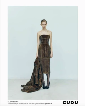 Model Bianca Szilágyi wearing a strapless brown leather midi dress and holding a matching coat against a light studio backdrop.