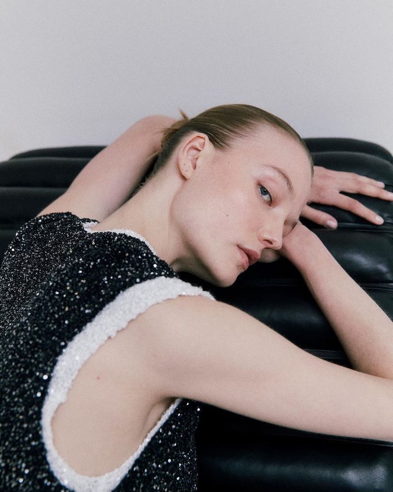 Woman in a black dress with white trim poses while reclining on a black leather couch in an indoor setting with a neutral background.