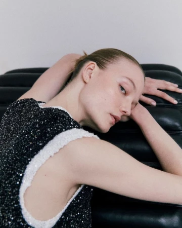 Woman in a black dress with white trim poses while reclining on a black leather couch in an indoor setting with a neutral background.