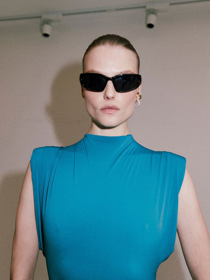 A woman poses indoors wearing a blue top and sunglasses, with sleek hair and a neutral expression, standing against a neutral wall.