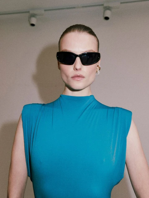 A woman poses indoors wearing a blue top and sunglasses, with sleek hair and a neutral expression, standing against a neutral wall.