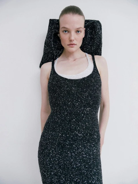 Elegant woman in a sparkling black dress with an architectural headpiece posing in a bright studio with soft, diffused lighting.