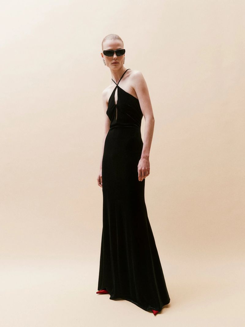 Elegant woman poses in a sleek black halter dress and sunglasses against a neutral backdrop, showcasing fashion and minimalist style.