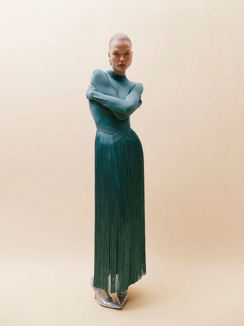 A fair-skinned woman in an aquamarine mock turtleneck and teal fringe skirt poses against a cream background in a fashion shot.