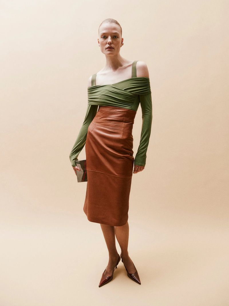 Adult female fashion model poses in a long-sleeve green draped top, brown leather skirt, brown heels and carrying a dark clutch purse.