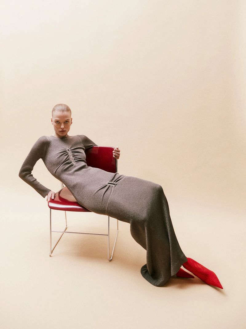 Fashion shot of a woman in a long gray dress and red boots sitting on a red chair against a light peach-colored background in a studio.