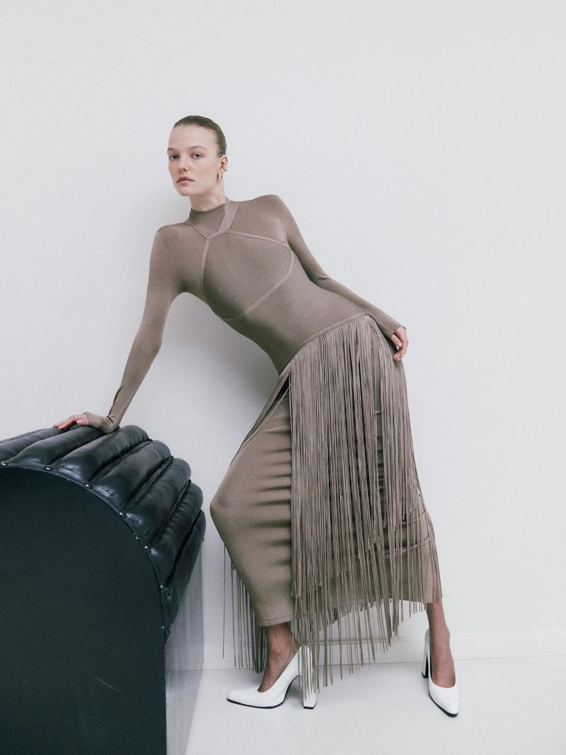 A slender woman poses in a stylish fringe dress and white heels, leaning against a black leather cushion in a minimalist studio setting.