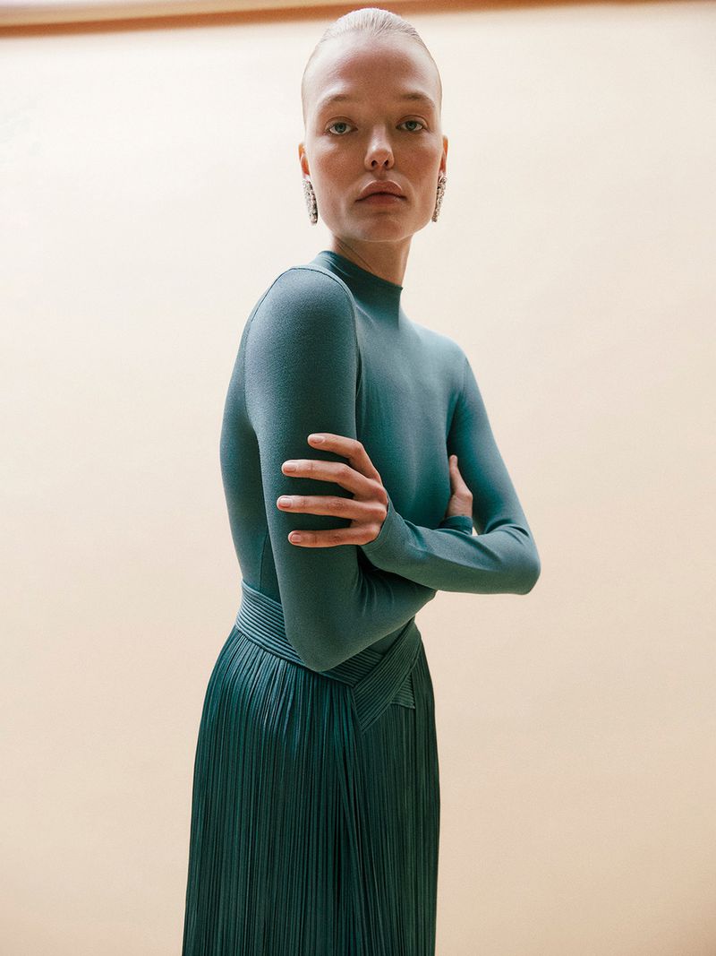Elegant woman in a teal top and pleated skirt posing in front of a neutral background, wearing large earrings.