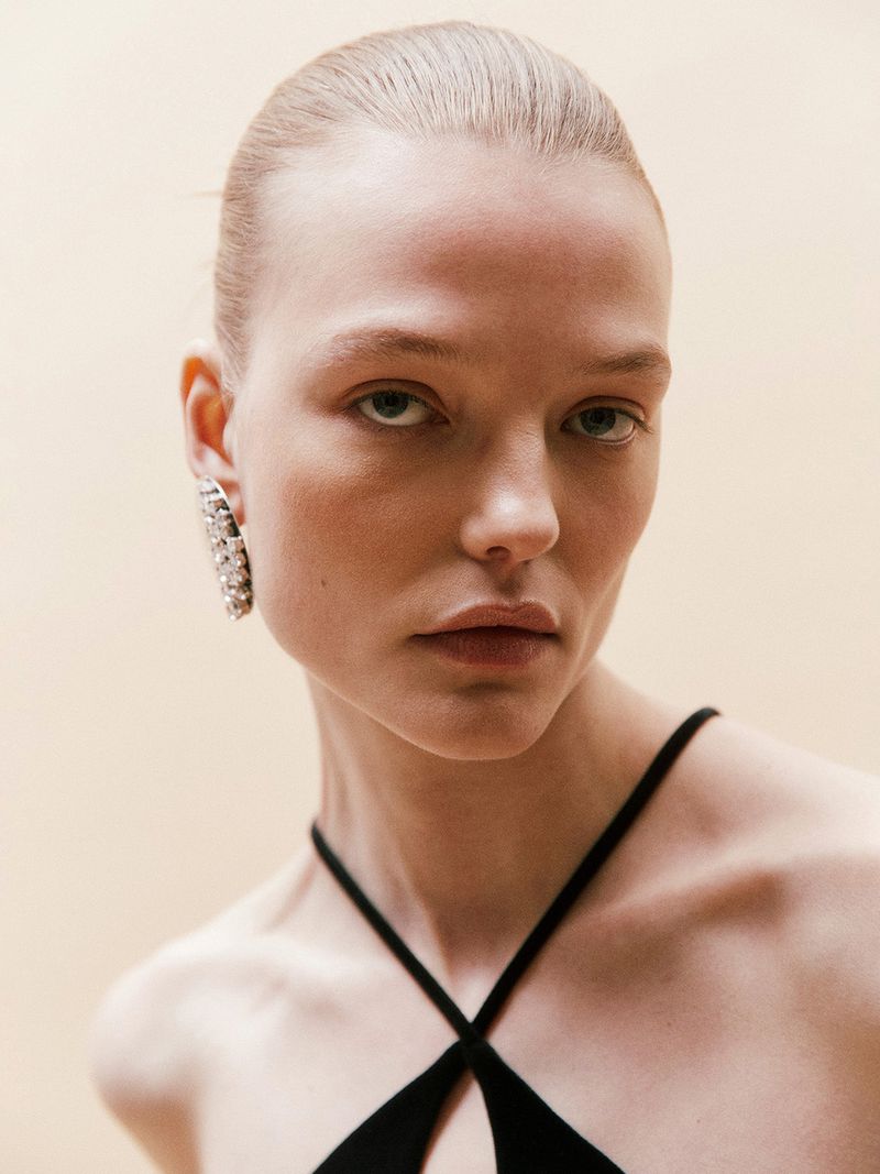 Close-up portrait of a fair-skinned adult woman with neutral facial expression, wearing a black top and large jeweled earring against a light background.