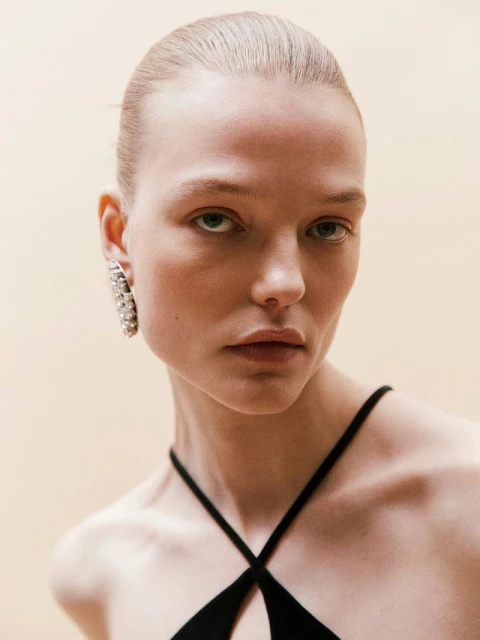 Close-up portrait of a fair-skinned adult woman with neutral facial expression, wearing a black top and large jeweled earring against a light background.