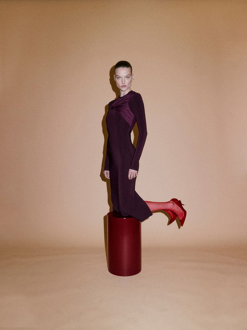 An adult female model poses on a red plinth in a burgundy dress and red high heels against a peach-colored backdrop in a fashion studio.