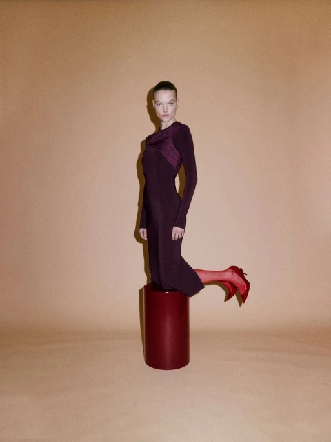 An adult female model poses on a red plinth in a burgundy dress and red high heels against a peach-colored backdrop in a fashion studio.