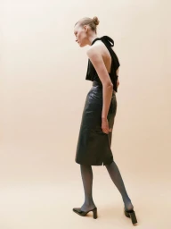 Model Roos Abels in profile wearing a black backless halter top and black leather midi skirt against a warm beige background.