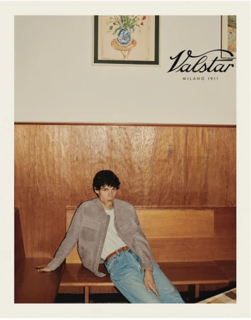 A stylish young man poses in a tan jacket and jeans in an indoor setting with wood paneling, captured in a medium shot.