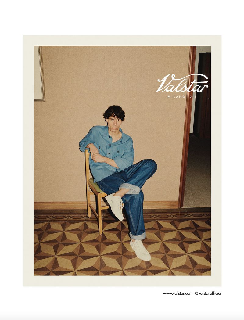 Stylish adult male poses on a chair indoors wearing a blue shirt and jeans with white sneakers, showcasing a casual fashion look.