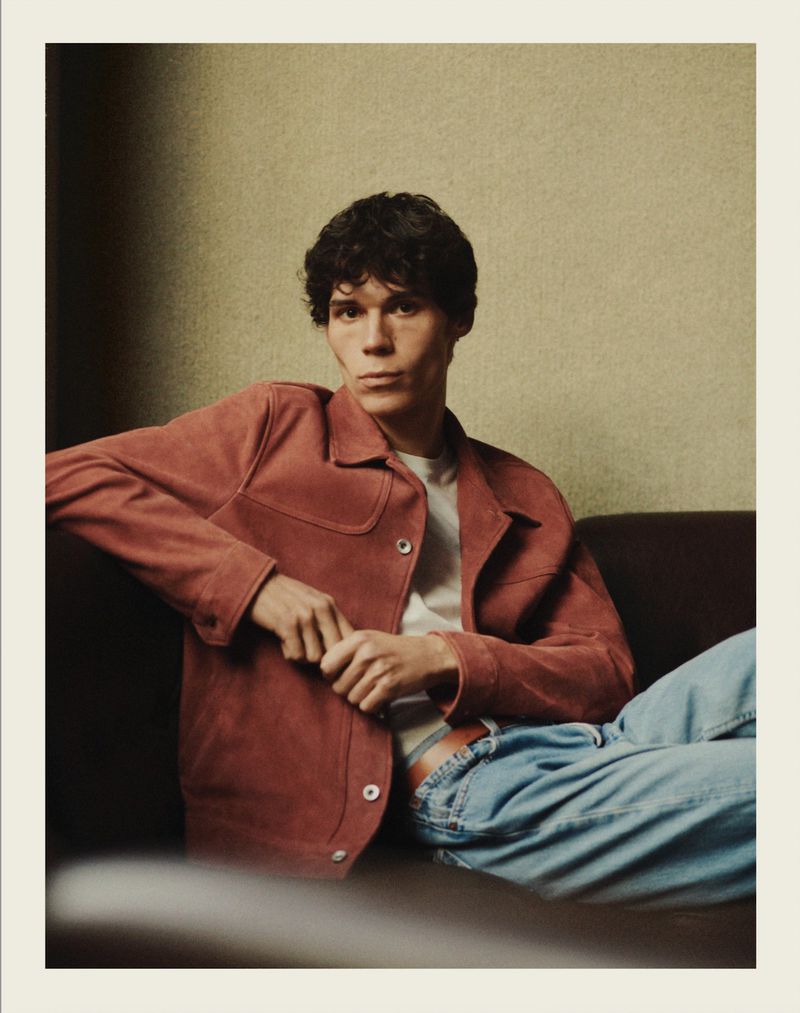 A young man with dark curly hair lounges on a sofa in casual apparel. He wears a brick-red jacket and light blue jeans, and looks at the camera.