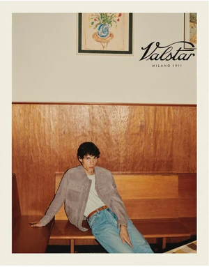 A young male model seated on a wooden bench wearing a light jacket, light t-shirt, and jeans, with Valstar branding visible.