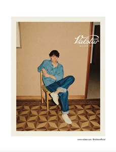 Model Tyler Goulden seated on a wooden chair wearing a blue overshirt and cuffed denim jeans on a geometric parquet floor.