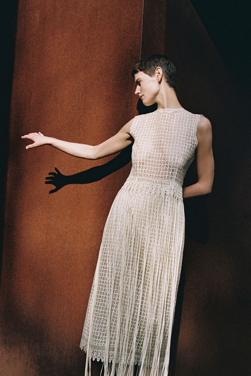 Adult woman in cream dress poses beside rust-colored column indoors with dark background, showing modern fashion style, beauty.