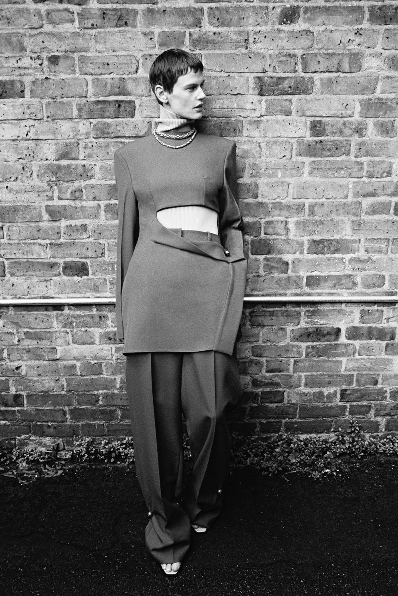 Monochrome fashion shot of a woman in a cutout top and wide-leg pants leaning against a brick wall in a full-length pose.