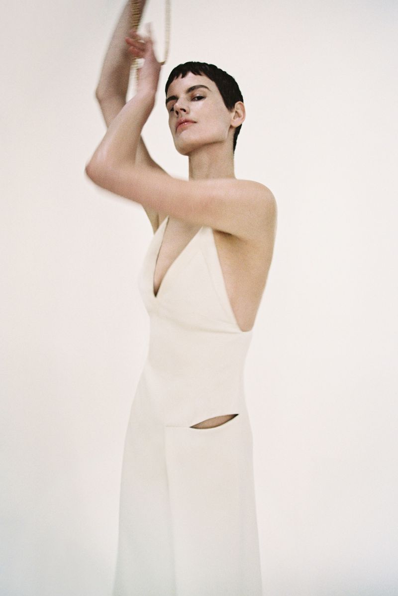 Woman in ivory halter top and wide-leg pants poses in a studio. She is fair-skinned with short dark hair, arms raised and hands near her face.