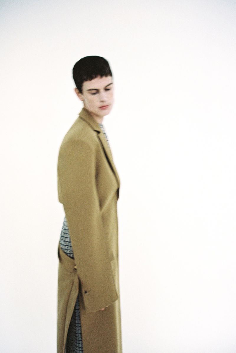 A woman with short, dark hair poses in a tan coat with a patterned dress underneath in a fashion studio setting with a white background.