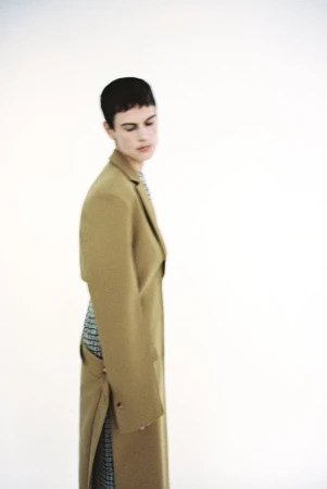 A woman with short, dark hair poses in a tan coat with a patterned dress underneath in a fashion studio setting with a white background.