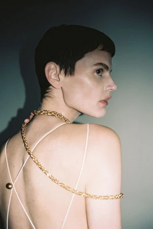 Close-up portrait of a female with short dark hair and a golden chain across her shoulder, looking away in a studio setting.
