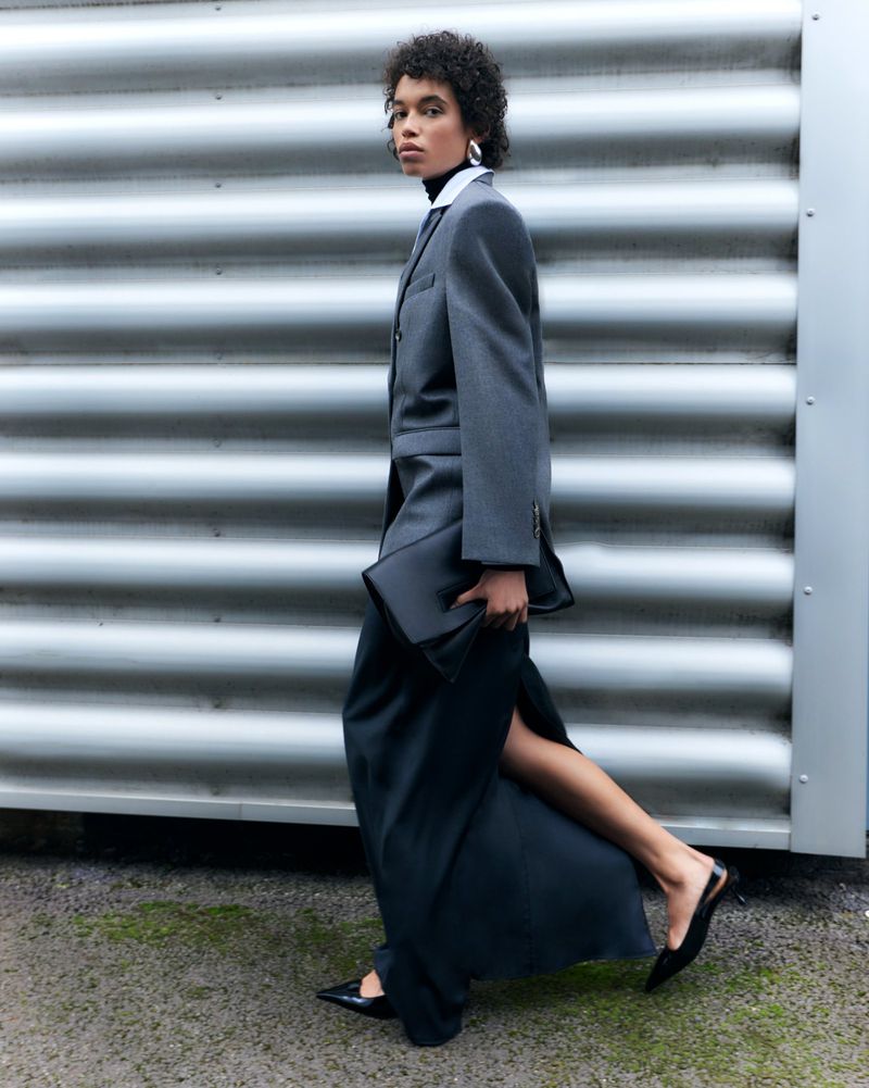 Fashionable woman in a gray suit and long black skirt walks against a textured silver wall with black handbag and shoes.