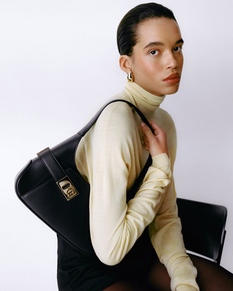 Model wearing a pale yellow turtleneck sweater carrying a black leather shoulder bag against a white studio background.