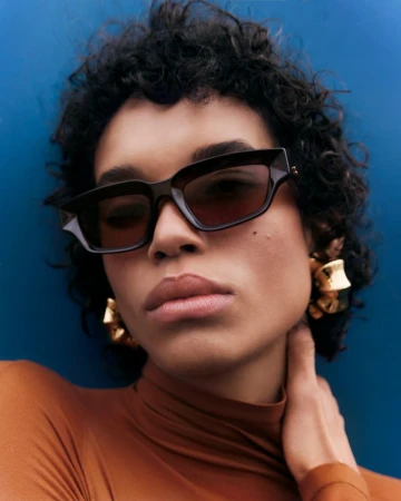 Close-up of a woman wearing sunglasses and gold statement earrings, posing in front of a dark blue wall, fashion style.
