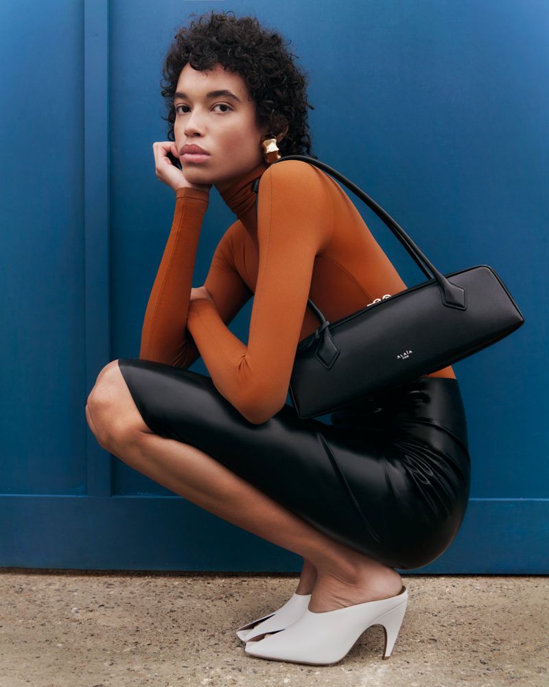 Woman in orange turtleneck and black skirt poses with a black handbag against a blue wall, showcasing fashion and style.