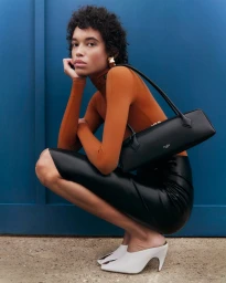 Model Lara Menezes crouching in an orange turtleneck, black leather skirt, and white mules with a black structured shoulder bag.