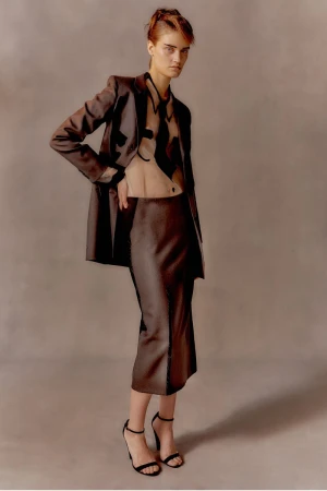 Model posing in a brown satin blazer, matching midi skirt, and sheer graphic shirt against a neutral backdrop.