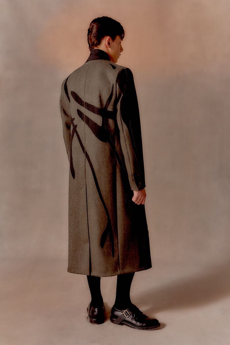 Back view of an adult male in a modern fashion coat with black accents, wearing black stockings and polished black shoes against a neutral backdrop.