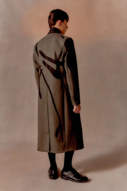 Back view of an adult male in a modern fashion coat with black accents, wearing black stockings and polished black shoes against a neutral backdrop.