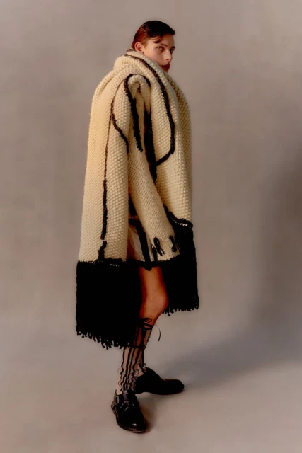 Fashion shot of a slender young adult posing in a cream-colored, knee-length dress paired with an oversized knit coat and black patent shoes.