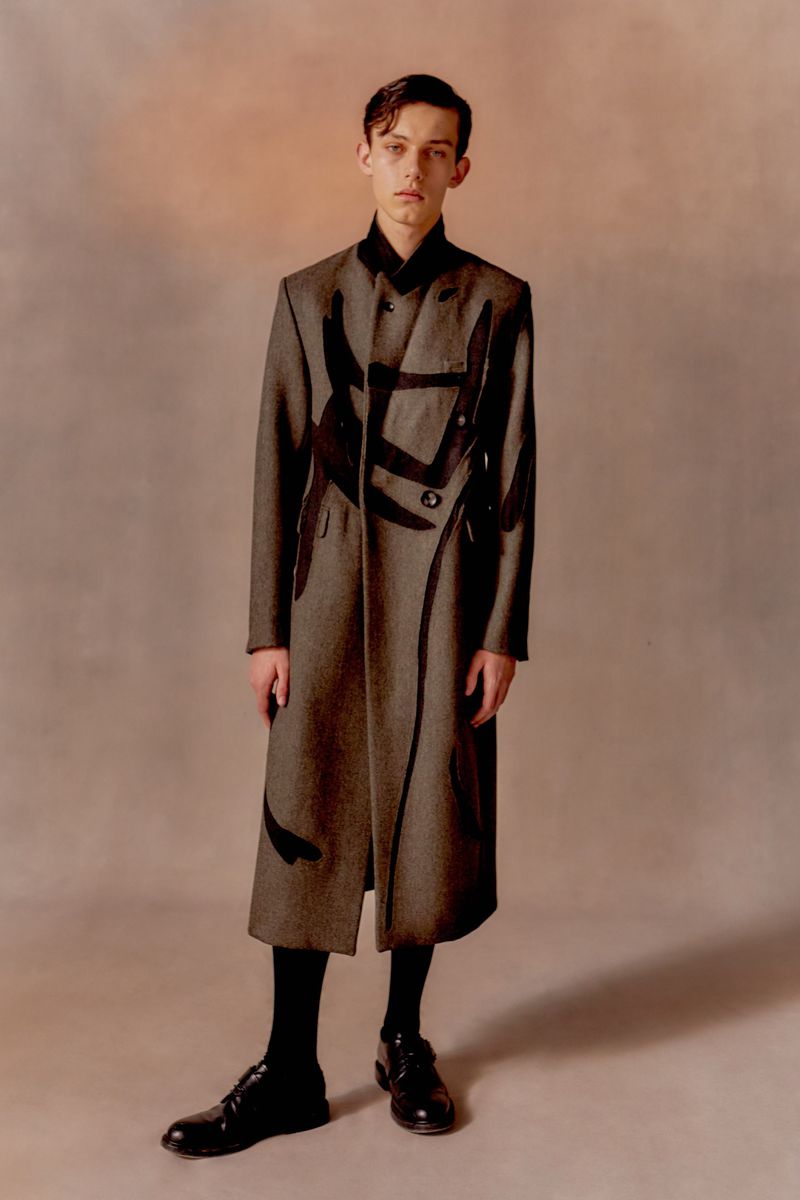 A young adult male stands in a full-length shot, wearing a gray double-breasted coat with dark graphics, black socks, and black dress shoes.