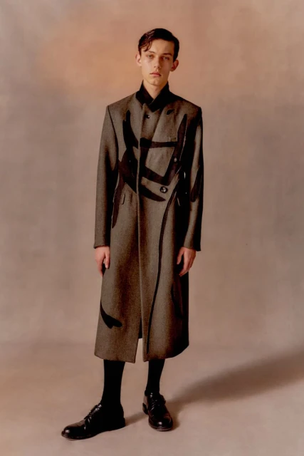A young adult male stands in a full-length shot, wearing a gray double-breasted coat with dark graphics, black socks, and black dress shoes.