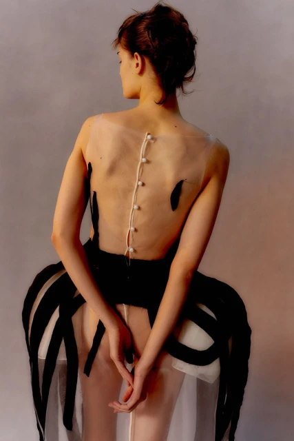 Back view of a female fashion model in a sheer white dress with black accents, featuring an open-back design with button details.