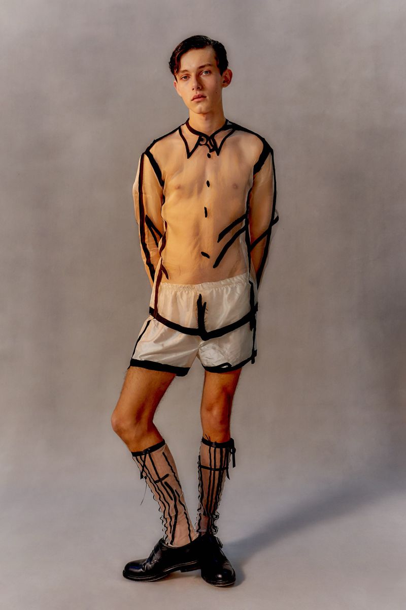 A slender young man strikes a pose in a see-through shirt and shorts, highlighted by black trim, standing in front of a neutral backdrop.