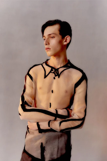 Portrait of a young adult man in a sheer black-outlined shirt looking to the left against a neutral background in eye-level shot.